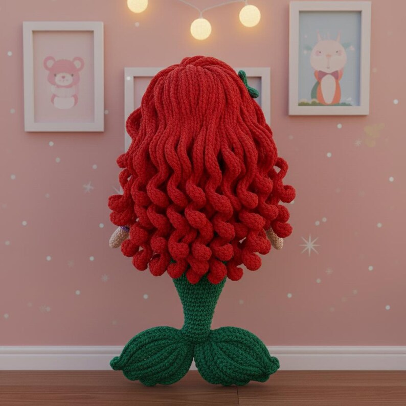 Op de afbeelding: Een handgemaakte zeemeerminnenpop met rood haar en een groene staart. Het haar van de pop is gestyled in grote, krullende golven. De staart van de zeemeermin is groen en heeft een waaiervormig ontwerp. De pop is gemaakt van garen en is waarschijnlijk een decoratief item.