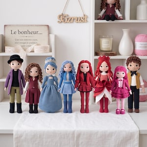 May include: A collection of eight handmade crochet dolls featuring diverse outfits and hairstyles. The dolls are arranged in a row, showcasing various colors including purple, red, blue, and pink. A sign with the text Le bonheur is visible.