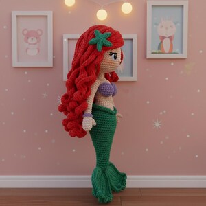 Op de afbeelding: Handgemaakte gehaakte pop van een zeemeermin met rood haar, een groene staart en een paarse top. De pop heeft een groene zeester in haar haar. De pop staat voor een roze muur met ingelijste foto's.