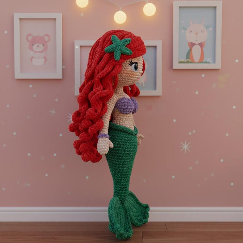 Op de afbeelding: Handgemaakte gehaakte pop van een zeemeermin met rood haar, een groene staart en een paarse top. De pop heeft een groene zeester in haar haar. De pop staat voor een roze muur met ingelijste foto's.
