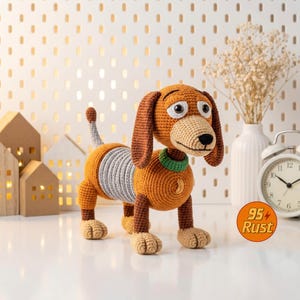 May include: A crocheted dog toy in shades of brown, tan, and gray, with a green collar. The toy has large, expressive eyes and a smiling face. The dog is standing on a white surface, with decorative houses and a clock in the background. The text "95 Rust" is in a circle.