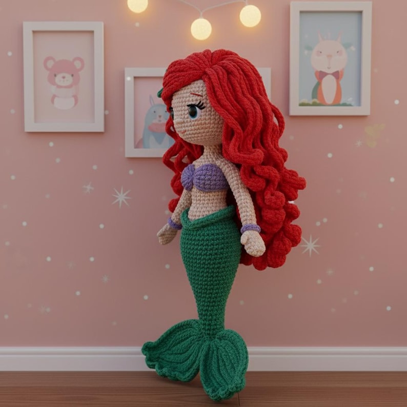Op de afbeelding: Handgemaakte gehaakte pop van een zeemeermin met felrood haar, een paarse top en een groene staart. De pop staat voor een roze muur met ingelijste foto's en decoratieve lampjes. De pop is ongeveer 30 cm hoog.