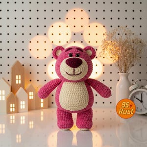 May include: A handmade pink crochet bear with a cream-colored belly and a dark pink nose and mouth. The bear has small ears and is standing upright. The background includes decorative lights, a white vase with dried flowers, and a small white alarm clock.
