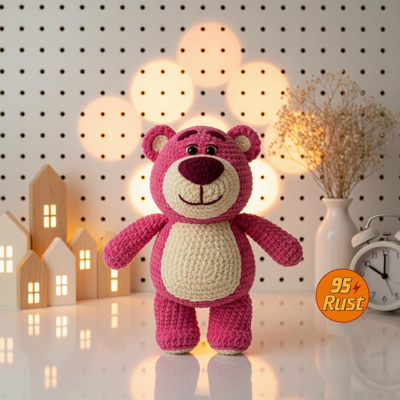 May include: A handmade pink crochet bear with a cream-colored belly and a dark pink nose and mouth. The bear has small ears and is standing upright. The background includes decorative lights, a white vase with dried flowers, and a small white alarm clock.
