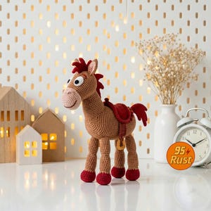 May include: A crocheted brown horse toy with a red saddle and red accents on its hooves and mane. The toy has a smiling face and is standing on a white surface. The background includes decorative houses, a vase with dried flowers, and a clock.