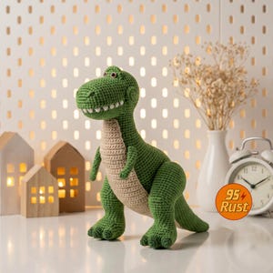 May include: A green and tan crocheted dinosaur toy with a friendly expression. The dinosaur has a detailed, textured appearance, with visible stitches and a smiling mouth. The toy is set on a white surface, with decorative elements in the background.