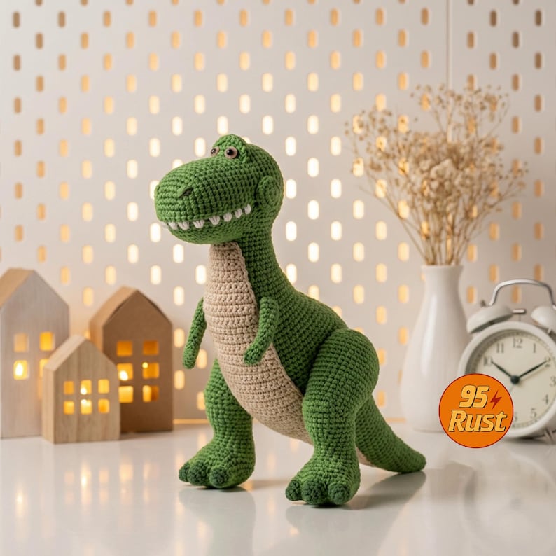 May include: A green and tan crocheted dinosaur toy with a friendly expression. The dinosaur has a detailed, textured appearance, with visible stitches and a smiling mouth. The toy is set on a white surface, with decorative elements in the background.