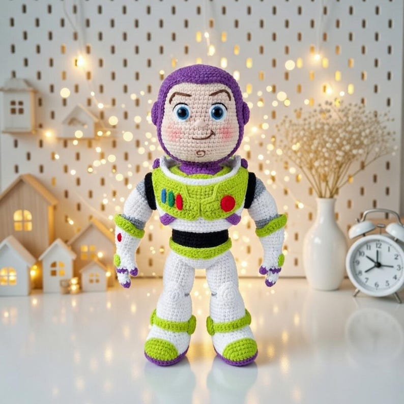 May include: A handmade crochet Buzz Lightyear doll, featuring a purple helmet, green and white spacesuit, and rosy cheeks. The doll is approximately 12 inches tall and stands on a white surface. The background includes decorative lights and miniature wooden houses.