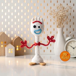 May include: A handmade crochet Forky doll, featuring a white body, red arms and a red unibrow. The doll has black eyes and a blue smile. The doll is standing on a white surface with a pegboard background and decorative houses.