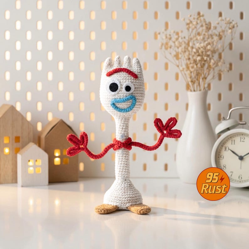 May include: A handmade crochet Forky doll, featuring a white body, red arms and a red unibrow. The doll has black eyes and a blue smile. The doll is standing on a white surface with a pegboard background and decorative houses.