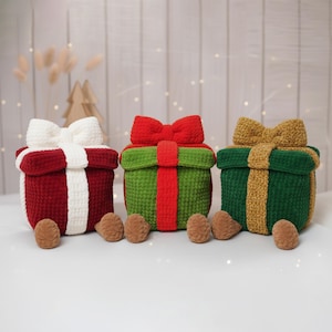 May include: Three crocheted gift boxes in red, green, and dark green, each with a contrasting bow and ribbon. The red box has a white bow, the green box has a red bow, and the dark green box has a gold bow. Each box has small brown feet.