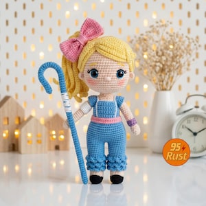 May include: A handmade crochet doll with blonde hair styled in a high ponytail with a pink bow. The doll wears a blue jumpsuit with a pink belt and holds a blue shepherd's crook. The doll has blue eyes and rosy cheeks.