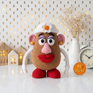 May include: A crocheted Mr. Potato Head doll with a brown body, white hat with a flower, pink ears, and red lips and shoes. The doll has white arms and is set against a white background with decorative elements.