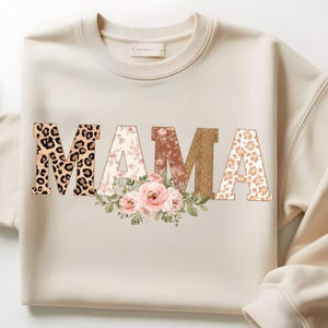 Peut inclure: Sweat-shirt crème avec le mot "MAMA" dans un mélange de motifs et de textures. Le "M" présente un imprimé léopard, le "A" a un motif floral, le deuxième "M" est doré pailleté et le dernier "A" a un motif floral. Un arrangement floral est centré sous le mot.