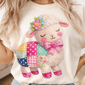 May include: A cream-coloured t-shirt with a charming lamb illustration. The lamb has a patchwork body in various colours and patterns, a pink bow, and a floral crown. The design is soft and detailed, ideal for a spring or Easter theme.