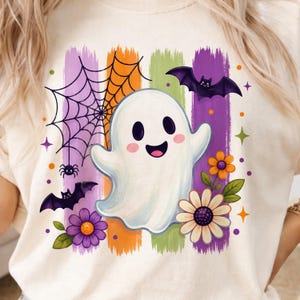 Könnte beinhalten: Ein cremefarbenes T-Shirt mit einer zentralen Grafik eines lächelnden weißen Geistes. Das Design umfasst violette, orangefarbene und grüne Pinselstrich-Streifen, ein schwarzes Spinnennetz mit Spinne, zwei violette Fledermäuse sowie orangefarbene und weiße Blumen.