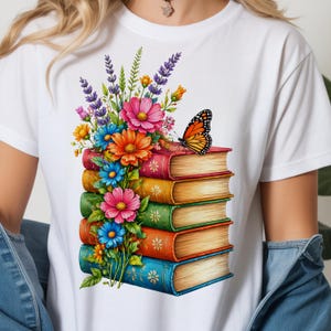 May include: White t-shirt featuring a colorful illustration of a stack of books adorned with flowers and a butterfly. The books have red, orange, green, and blue covers. The flowers include pink, blue, orange, and yellow blooms.