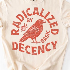 Op de afbeelding: Crèmekleurig T-shirt met een verweerde rode afbeelding. De afbeelding toont een ravenillustratie omcirkeld door de woorden "RADICALIZED BY BASIC DECENCY". Een takje bladeren staat naast de raaf. Het shirt heeft opgerolde mouwen.