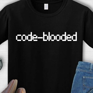 Puede incluir: Camiseta negra con el texto blanco "code-blooded" en una fuente pixelada. La camiseta se combina con vaqueros azules, botas de cuero marrones y gafas de sol.