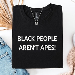 May include: Black t-shirt with the text "BLACK PEOPLE AREN'T APES!" in white. A silver necklace with a sunburst pendant is on the shirt. The shirt is folded on a white surface, next to blue jeans and white shoes.
