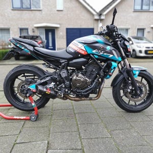 以下が含まれることがあります： 黒とティールのヤマハMT-07オートバイがレンガの表面に駐車されています。オートバイは黒いホイール、黒いシート、ティールアクセントを備えています。オートバイは赤いスタンドにあります。背景には家と車が含まれています。