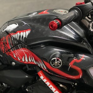 以下が含まれることがあります： 赤と白のモンスターの顔のグラフィックデザインが施された黒いオートバイの燃料タンク。オートバイは、赤いアクセントとヤマハのロゴが付いた黒いハンドルバーを備えています。