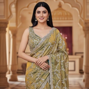 May include: A woman wearing a gold-colored sari with silver embroidery. The sleeveless blouse and sari are adorned with intricate floral patterns and a silver border. The background features an ornate architectural design.