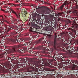 May include: A burgundy saree with intricate floral embroidery and a matching blouse. The saree features a detailed border and is adorned with sparkling embellishments. A gold-colored decorative item is placed on the saree.