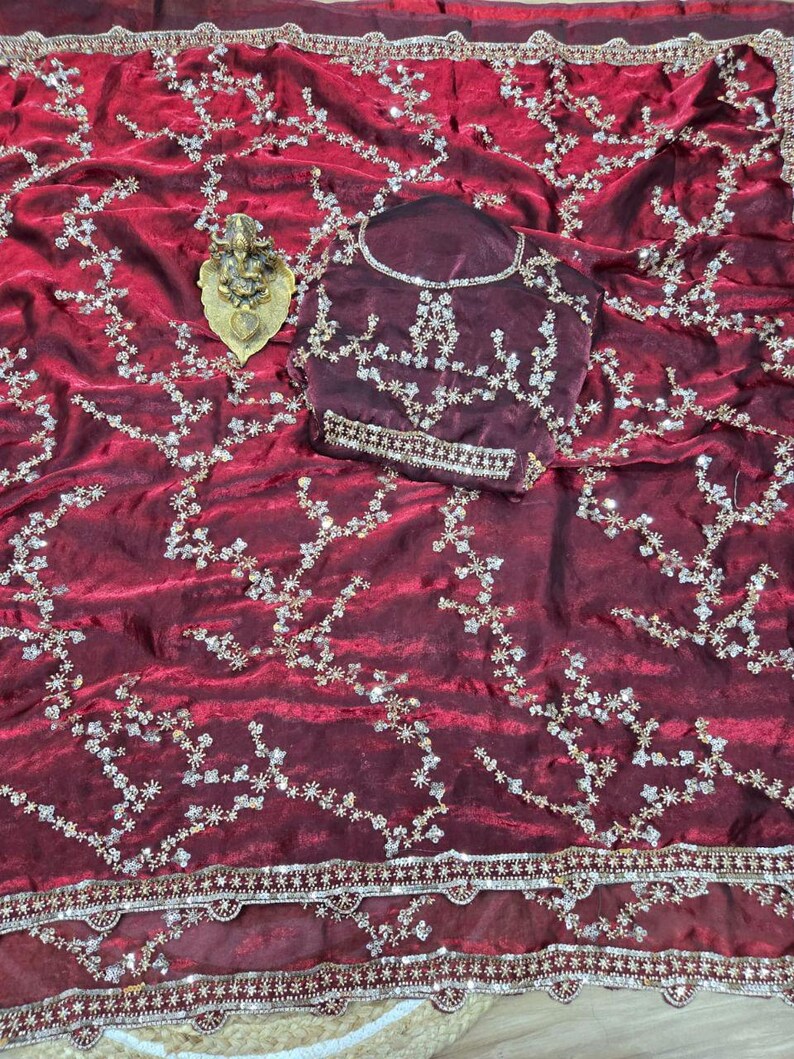 May include: A burgundy saree with intricate floral embroidery and a matching blouse. The saree features a detailed border and is adorned with sparkling embellishments. A gold-colored decorative item is placed on the saree.