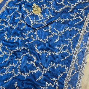 May include: Royal blue sari with intricate silver embroidery. The fabric is draped, showcasing a detailed floral pattern. A small gold statue rests on the fabric. The sari's border features a decorative silver trim, adding a touch of elegance.