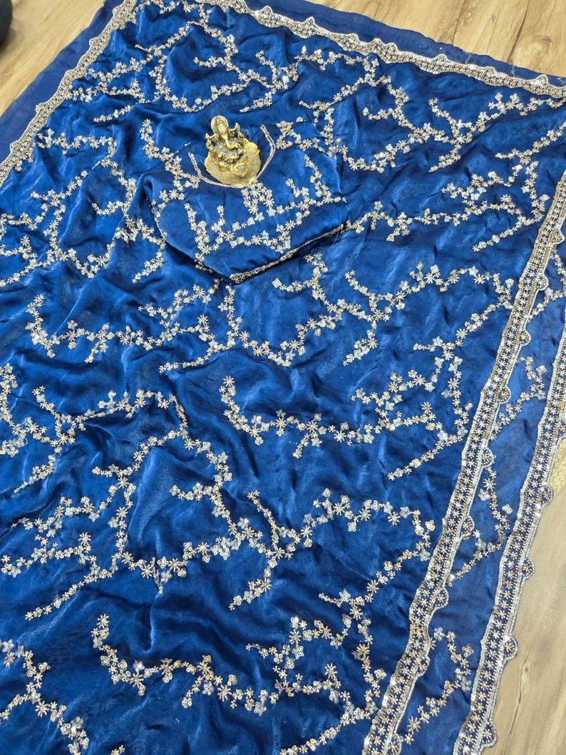 May include: Royal blue sari with intricate silver embroidery. The fabric is draped, showcasing a detailed floral pattern. A small gold statue rests on the fabric. The sari's border features a decorative silver trim, adding a touch of elegance.