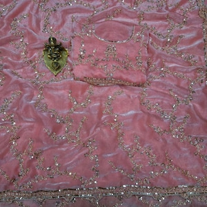 May include: A pink velvet saree with intricate gold embroidery and sequin embellishments. The saree includes a matching blouse piece, also adorned with gold embroidery. A gold-colored decorative item is placed on the saree.