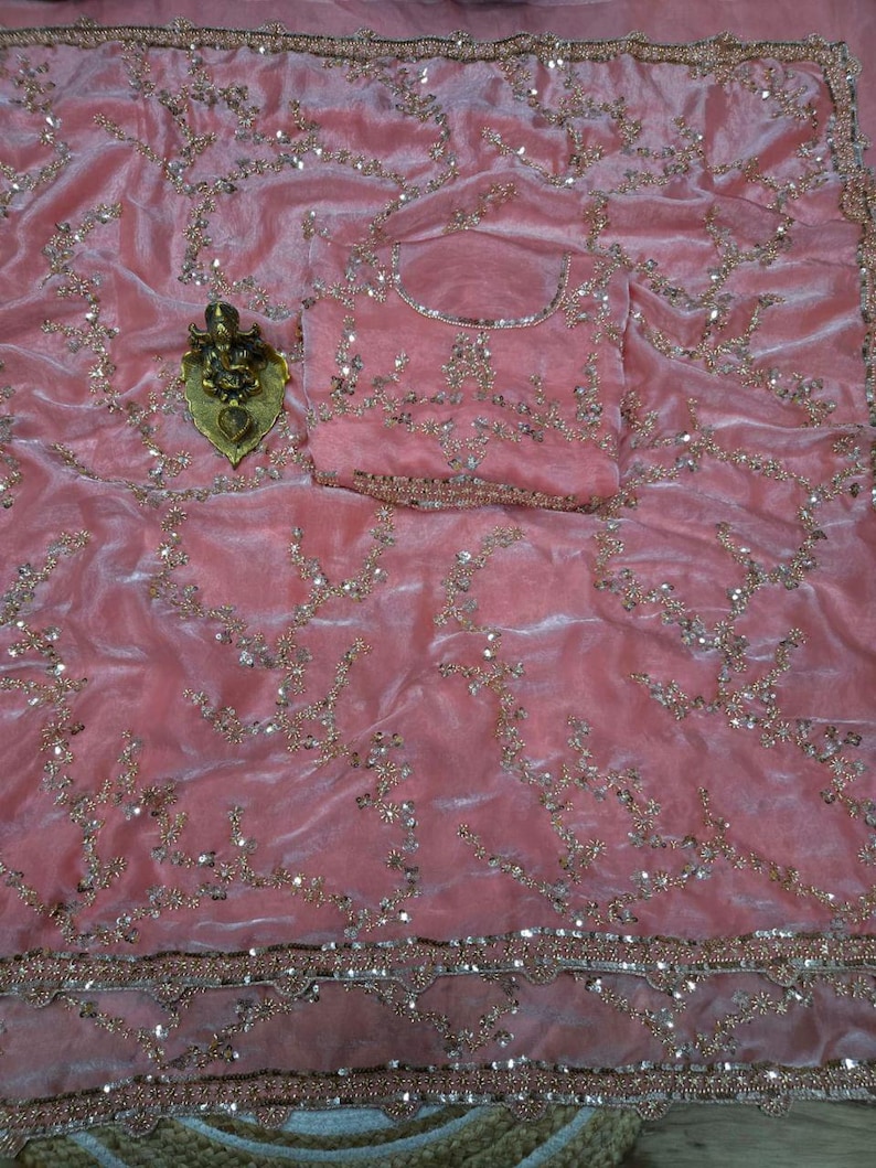 May include: A pink velvet saree with intricate gold embroidery and sequin embellishments. The saree includes a matching blouse piece, also adorned with gold embroidery. A gold-colored decorative item is placed on the saree.