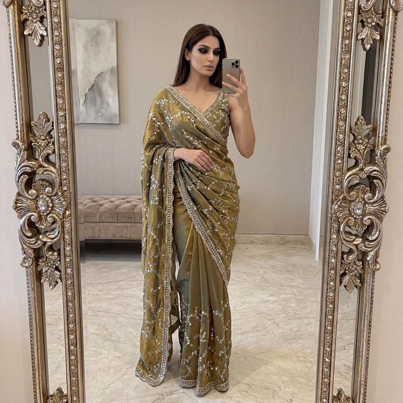 May include: A gold-colored sari with intricate silver embroidery and embellishments. The sari features a V-neck design and a draped style, reflecting a traditional South Asian garment. The fabric has a subtle sheen, adding to its elegance.