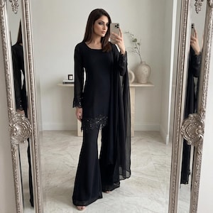 Puede incluir: Conjunto negro de manga larga con pantalón acampanado. Las mangas y el pantalón están adornados con delicados bordados florales. Una bufanda transparente a juego completa el conjunto. El conjunto se refleja en un espejo ornamentado.