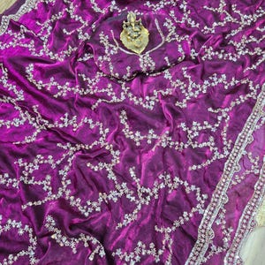 May include: A vibrant magenta saree with intricate silver floral embroidery. The fabric drapes elegantly, showcasing a detailed border and a matching blouse piece. A golden deity statue rests on the blouse piece.