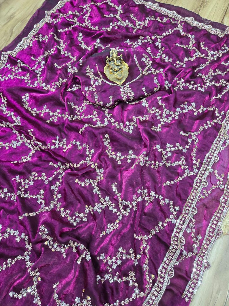 May include: A vibrant magenta saree with intricate silver floral embroidery. The fabric drapes elegantly, showcasing a detailed border and a matching blouse piece. A golden deity statue rests on the blouse piece.