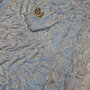 May include: A light blue sari with intricate silver embroidery and embellishments. The fabric has a shimmering, textured appearance. A small, gold-colored statue rests on the fabric. The sari's border is detailed with a scalloped edge.