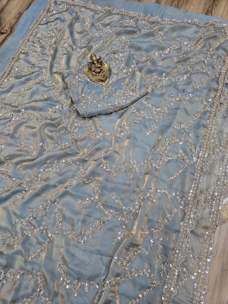 May include: A light blue sari with intricate silver embroidery and embellishments. The fabric has a shimmering, textured appearance. A small, gold-colored statue rests on the fabric. The sari's border is detailed with a scalloped edge.