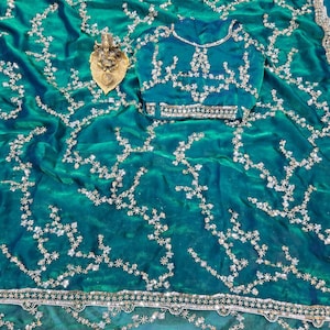 May include: A teal saree with intricate silver embroidery and a matching blouse. The saree features a detailed floral pattern and a decorative border. A small gold statue rests on the saree.