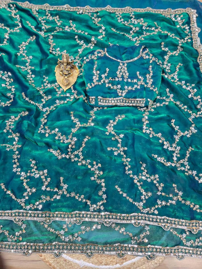 May include: A teal saree with intricate silver embroidery and a matching blouse. The saree features a detailed floral pattern and a decorative border. A small gold statue rests on the saree.