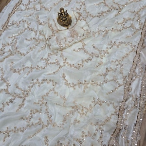 May include: Ivory-colored fabric with intricate gold sequin detailing. The fabric is draped, showcasing a flowing design. A gold-colored statue rests on the fabric. The edges are trimmed with a gold border, adding a touch of elegance. The overall theme is luxury and textiles.