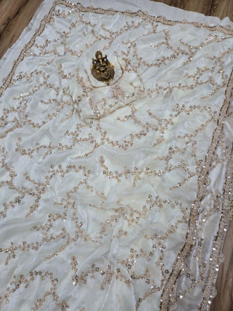 May include: Ivory-colored fabric with intricate gold sequin detailing. The fabric is draped, showcasing a flowing design. A gold-colored statue rests on the fabric. The edges are trimmed with a gold border, adding a touch of elegance. The overall theme is luxury and textiles.