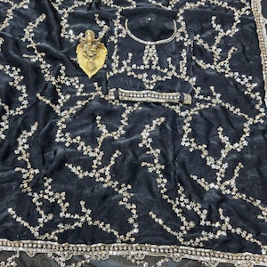 May include: A black velvet saree with intricate floral embroidery in gold and silver. The saree features a matching blouse and a decorative border. A small gold statue rests on the fabric.