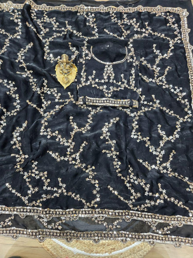 May include: A black velvet saree with intricate floral embroidery in gold and silver. The saree features a matching blouse and a decorative border. A small gold statue rests on the fabric.