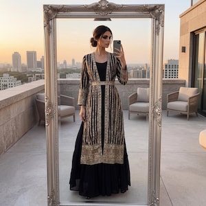May include: A woman in a black dress and a long, open jacket is reflected in a full-length mirror. The jacket is black with vertical gold stripes and gold embroidery. The mirror has a decorative silver frame. The background shows a city skyline at sunset.