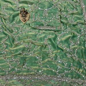 May include: A green sari with intricate silver and gold embroidery. The fabric has a shimmering texture and is adorned with floral and geometric patterns. A matching blouse piece is included. A small gold-colored statue is placed on the sari.