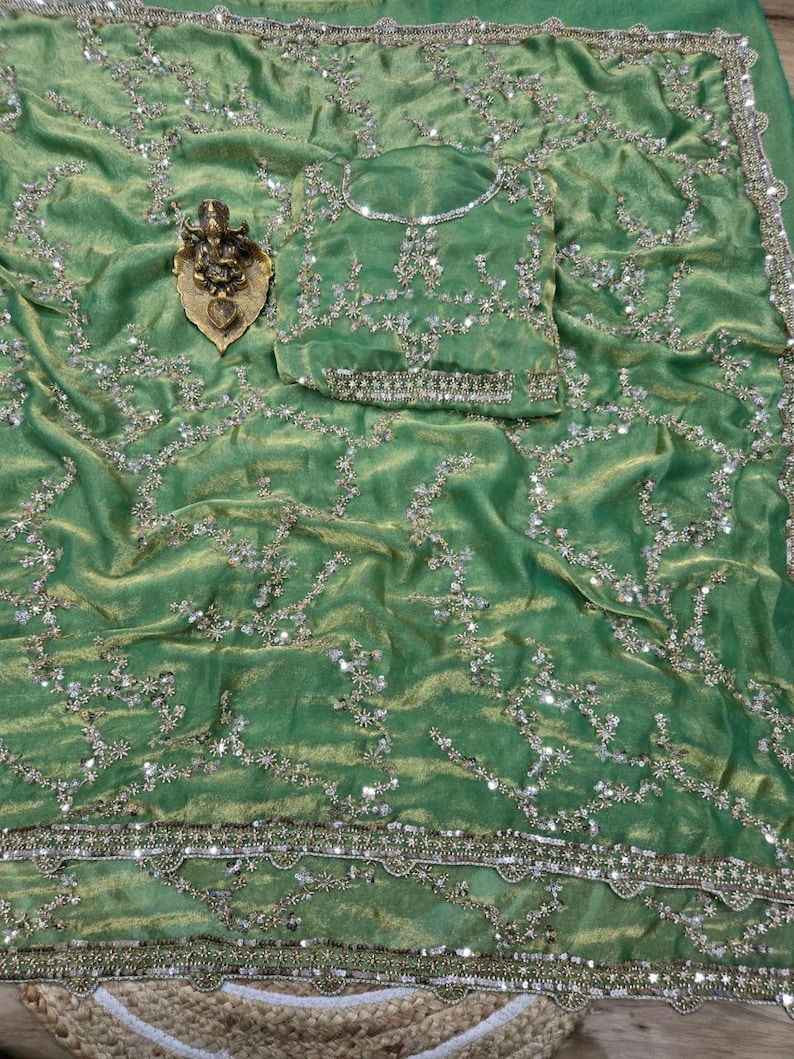 May include: A green sari with intricate silver and gold embroidery. The fabric has a shimmering texture and is adorned with floral and geometric patterns. A matching blouse piece is included. A small gold-colored statue is placed on the sari.