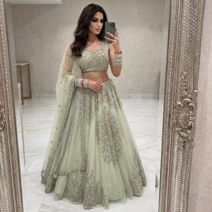 Puede incluir: Un conjunto lehenga choli verde claro adornado. El conjunto incluye un top corto con cuello en V, una falda larga y una dupatta transparente. La falda y el top están adornados con intrincados bordados plateados. El atuendo se captura en un espejo.