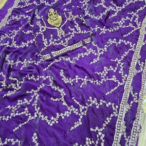 May include: A vibrant purple saree with intricate silver embroidery. The fabric is draped, showcasing a flowing design. The saree features a detailed border and a decorative gold-colored centerpiece.